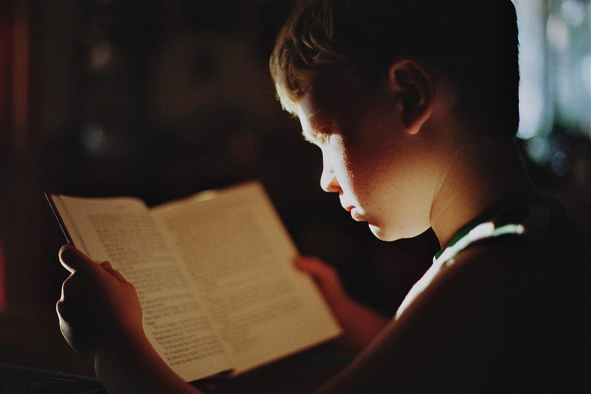 A child reading an open book by lamplight, face lit by the page.